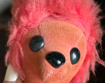 May include: A vintage pink plush toy head with black button eyes and a black nose. The toy has pink fluffy hair and two small, tan fabric horns. The toy's neck is attached to a decorative collar with gold and yellow trim.