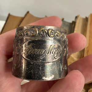 Puede incluir: Un servilletero plateado con el nombre "Anna May" grabado en escritura cursiva. El anillo tiene un dise&ntilde;o floral decorativo en el borde superior. El anillo est&aacute; en una mano.