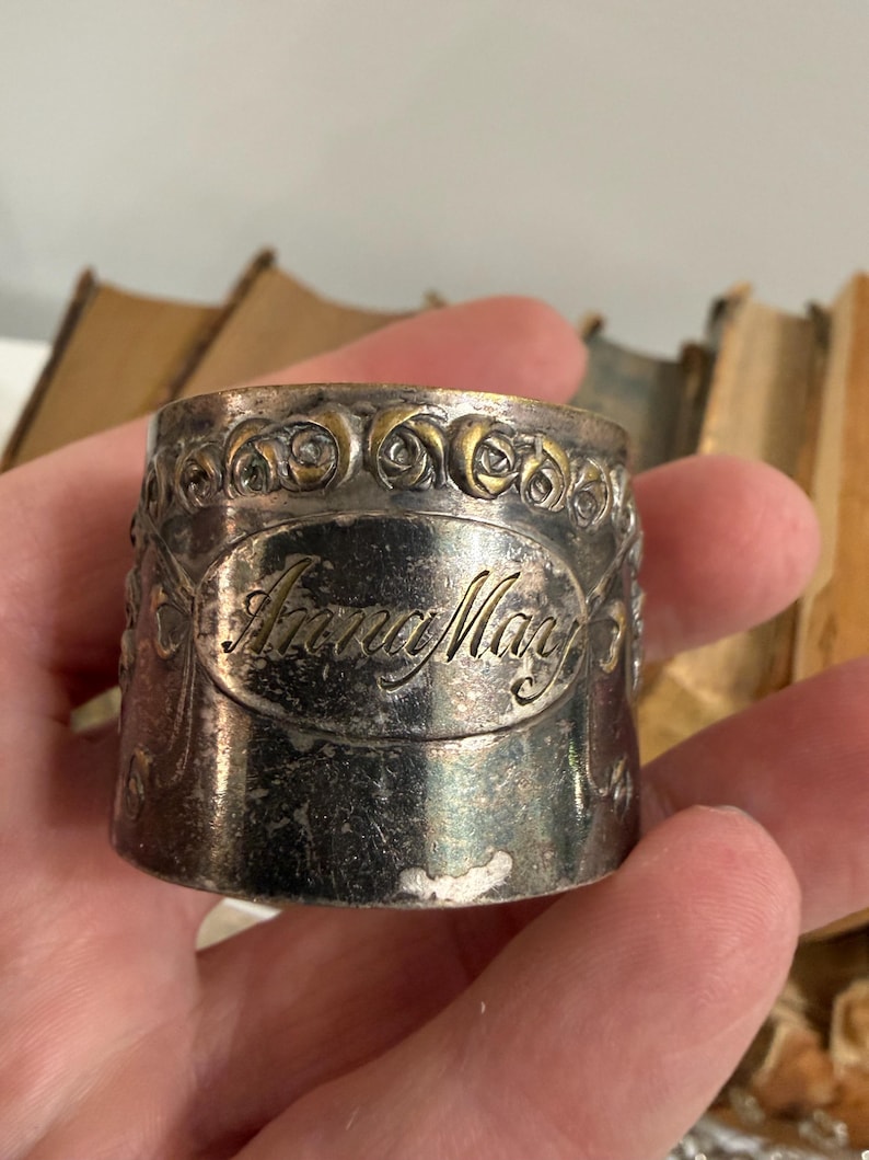 Puede incluir: Un servilletero plateado con el nombre "Anna May" grabado en escritura cursiva. El anillo tiene un dise&ntilde;o floral decorativo en el borde superior. El anillo est&aacute; en una mano.