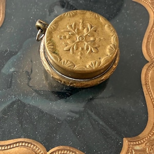 May include: A small, round, antique brass container with a hinged lid. The lid is decorated with an embossed floral design and a scalloped edge. A small loop is attached to the side for hanging. The container sits on a dark surface.