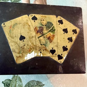 May include: A black rectangular box with playing card designs on the lid. The cards are Ace of Spades, King of Spades, and Ten of Spades. The cards have a vintage, distressed look.