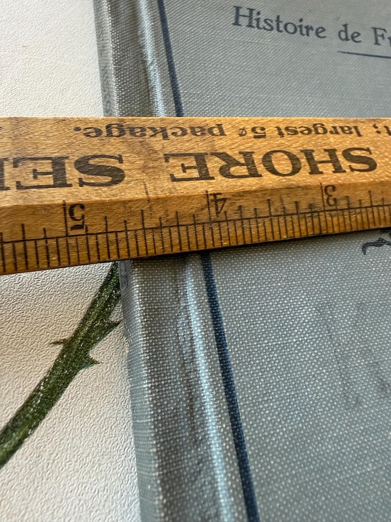 Puede incluir: Una regla de madera con marcas negras y el texto "SHORE SEI" se coloca sobre un libro azul claro. El libro tiene el t&iacute;tulo "Histoire de Fr" impreso en la portada. La regla mide hasta 12,7 cm.