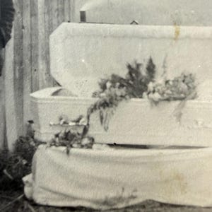 May include: Black and white photograph of three stacked, white caskets. The top casket is open, revealing a floral arrangement. The caskets are draped with white fabric. The image is taken outdoors.