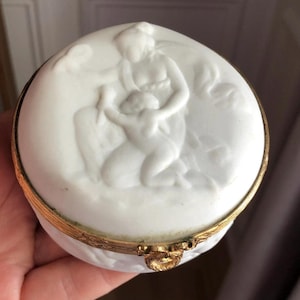 May include: A white porcelain trinket box with a gold-colored metal rim and clasp. The lid features a bas-relief sculpture of a woman and child. The box is round and appears to be vintage.