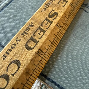 Puede incluir: Una regla de madera vintage con marcas negras y las palabras "SEED" y "MORE" impresas. La regla est&aacute; en &aacute;ngulo sobre una superficie azul estampada, con el texto "Ask Your" visible.