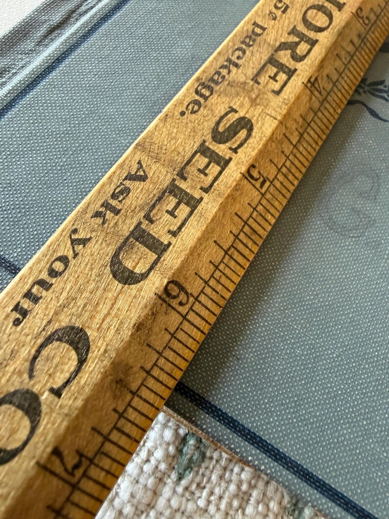 Puede incluir: Una regla de madera vintage con marcas negras y las palabras "SEED" y "MORE" impresas. La regla est&aacute; en &aacute;ngulo sobre una superficie azul estampada, con el texto "Ask Your" visible.