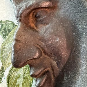 May include: Close-up of a dark brown, cast metal crescent moon with a face. The moon has a textured surface and a detailed face with a closed eye, a prominent nose, and a smiling mouth. The background has a floral design.