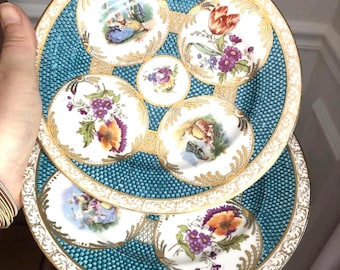 Könnte beinhalten: Zwei runde Zierteller mit türkisfarbenem, strukturiertem Hintergrund und goldenen Filigranrändern. Die Teller zeigen mehrere kreisförmige weiße Einsätze, die mit bunten Blumenarrangements und detaillierten Szenen von Figuren in historischer Kleidung verziert sind.