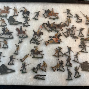 May include: A collection of antique toy soldiers and cannons, arranged on a white surface within a black framed display. The figures depict soldiers on foot and horseback, some holding flags, in various poses. The color palette includes shades of green, brown, and gold.