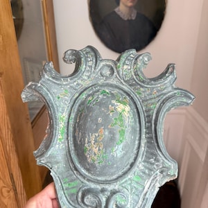 May include: An antique, ornate, gray-green wall plaque with an oval center and decorative scrollwork. The paint is distressed, revealing hints of green and other colors. The plaque is held in a hand.