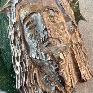 May include: A metallic, gold-toned relief plaque depicting the face of Jesus Christ with a crown of thorns. The sculpture features textured, carved details on the hair and beard, with a weathered, aged finish that highlights the facial features.