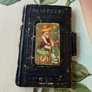 May include: A black leather-bound notebook with a worn cover. The front cover features a small image of a girl in a straw hat watering a rose bush. The text "PARCHMENT" is embossed on the cover. The text "NOTES" is embossed on the bottom of the cover.
