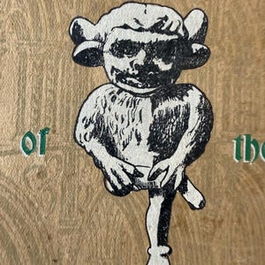 May include: A black and white illustration of a mythical creature with large ears and a human-like face. The creature is holding a small object and has a single leg. The background is a light brown wood grain.