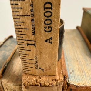 Puede incluir: Primer plano de una regla de madera vintage, marcada en pulgadas, apoyada en el lomo de un libro antiguo. La regla est&aacute; marcada "Made in U.S.A." y el texto "A GOOD R" es visible. El libro tiene una cubierta desgastada y texturizada.