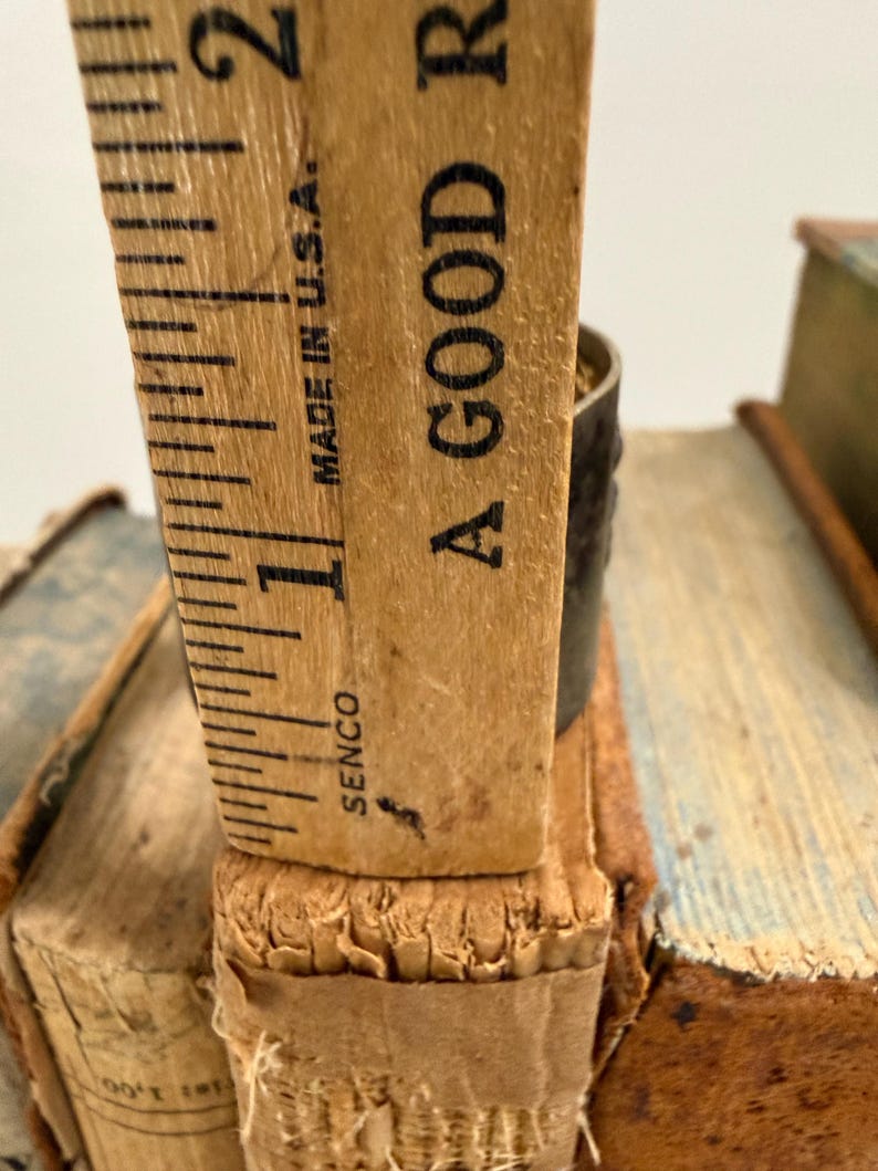 Puede incluir: Primer plano de una regla de madera vintage, marcada en pulgadas, apoyada en el lomo de un libro antiguo. La regla est&aacute; marcada "Made in U.S.A." y el texto "A GOOD R" es visible. El libro tiene una cubierta desgastada y texturizada.