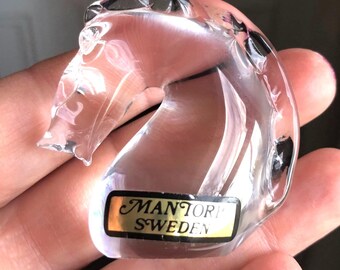 May include: A clear glass sculpture shaped like a stylized horse head. The piece features smooth, curved lines and a reflective surface. A rectangular gold-colored sticker is attached to the base, featuring the black text MANTORP SWEDEN.