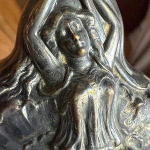 May include: Close-up of a bronze-colored sculpture featuring a woman with flowing hair and arms raised. The sculpture has intricate details and a classical art style. The woman's face is visible, and the sculpture appears to be part of a larger piece.