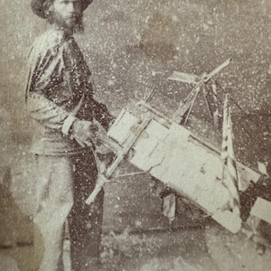 May include: Sepia-toned photograph of a man with a beard and hat, holding a complex mechanical device. The device appears to be made of wood and metal, with various levers and a rectangular box. The man is wearing a long-sleeved shirt and pants.