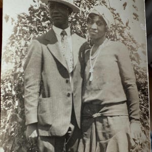 May include: A vintage black and white photograph of a couple posing outdoors. The man wears a light-colored suit, tie, and a fedora hat. The woman is in a dress and head covering, with a necklace. They stand in front of foliage.
