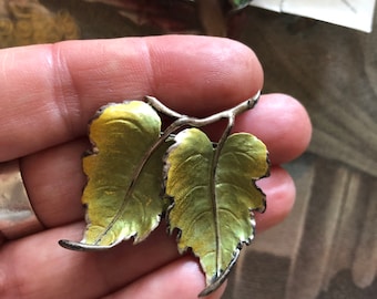 May include: A silver-toned brooch and matching earrings featuring a leaf design. The leaves are painted with shades of green and yellow, with a detailed, textured appearance. The brooch has two leaves, while the earrings each have a single leaf.