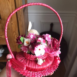 Puede incluir: Una cesta de sat&eacute;n rosa con asa, llena de flores de seda rosa, blanca y roja. Un peque&ntilde;o conejito blanco asoma desde el centro de la cesta.