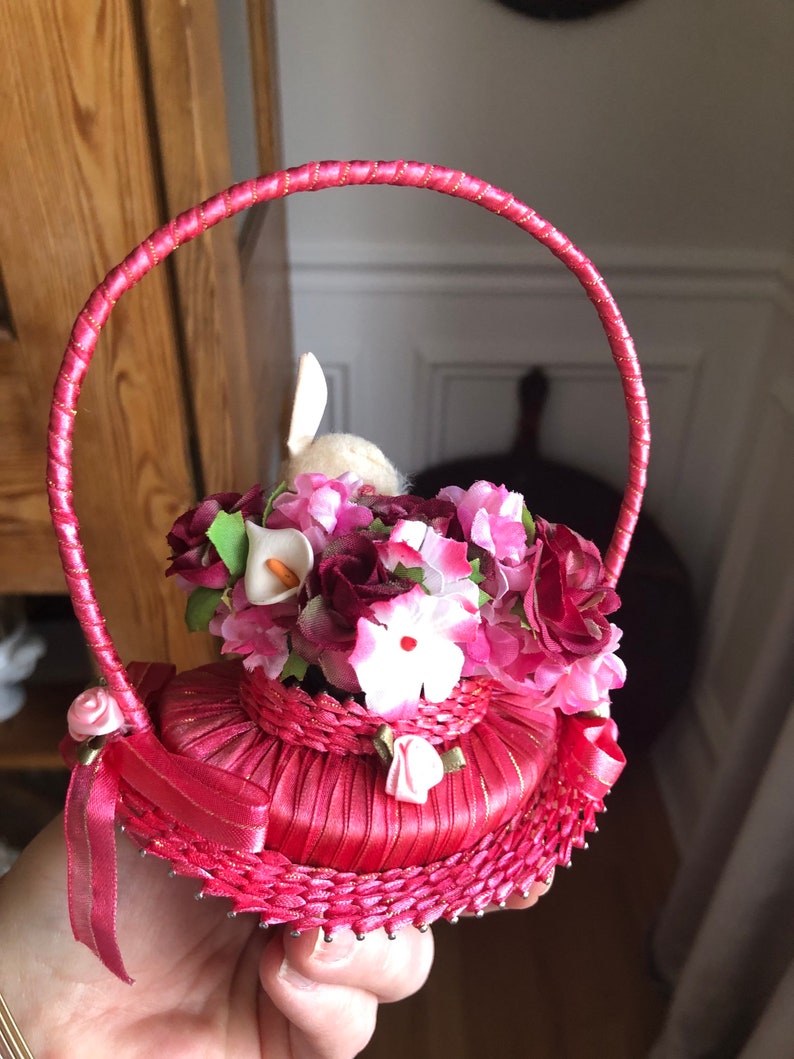 Puede incluir: Una cesta de sat&eacute;n rosa con asa, llena de flores de seda rosa, blanca y roja. Un peque&ntilde;o conejito blanco asoma desde el centro de la cesta.