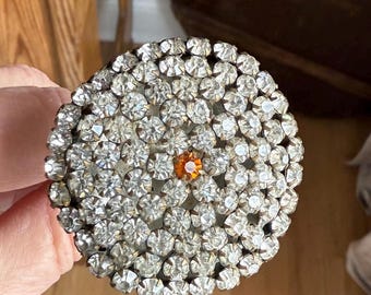 May include: A round brooch with a large cluster of clear rhinestones and a single orange rhinestone in the center.