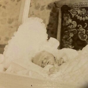 May include: A black and white photo of a baby sleeping in a crib. The baby is wearing a white dress and has a white blanket over them. The crib is made of wood and has a white fabric lining.