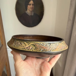 May include: A dark brown ceramic bowl with a decorative rim featuring a grape and leaf design in shades of green and gold. The bowl is oval-shaped and held in a hand.