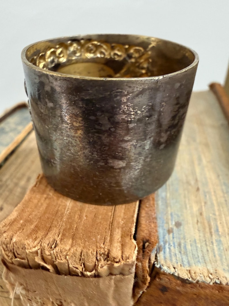 Puede incluir: Una peque&ntilde;a copa de plata antigua con un borde dorado decorativo y ornamentado. La copa es cil&iacute;ndrica y se apoya en una pila de libros viejos y desgastados. La superficie de la copa muestra signos de edad y uso.