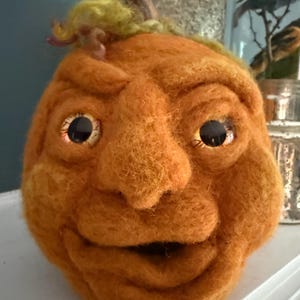 May include: A needle-felted orange pumpkin sculpture with a textured surface, featuring a brown stem, yellow fiber accents on top, and large, round, dark-pupiled eyes set into a sculpted face with a wide, open mouth and prominent nose.