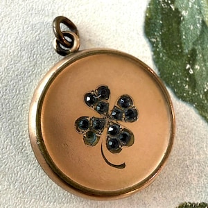 May include: A round, gold-toned metal locket pendant featuring a centered four-leaf clover motif inlaid with small, dark, faceted stones. The locket has a smooth, matte finish and includes a small circular jump ring at the top for attachment.