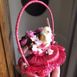 Puede incluir: Una cesta de Pascua de tela rosa con un conejito blanco y flores rosas y blancas. La cesta tiene una cinta rosa y un borde dorado.