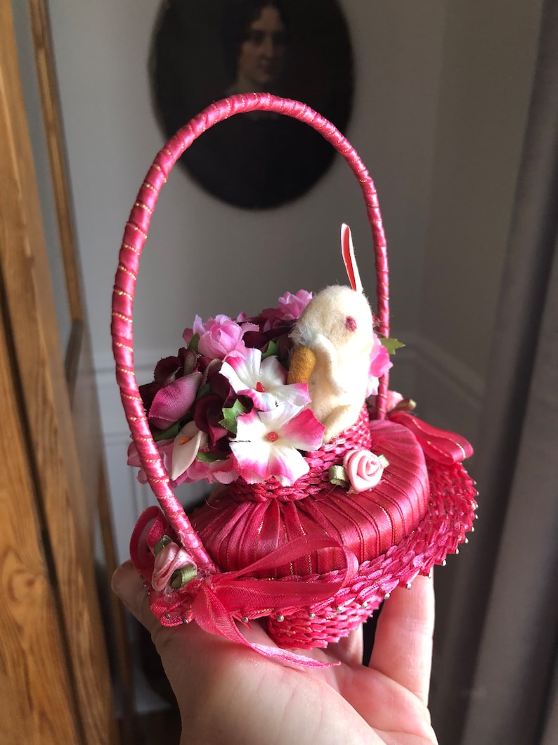 Puede incluir: Una cesta de Pascua de tela rosa con un conejito blanco y flores rosas y blancas. La cesta tiene una cinta rosa y un borde dorado.