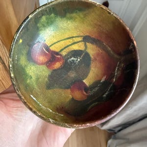 May include: A small, round wooden bowl with a hand-painted design of cherries. The bowl's interior features a vibrant color palette of red, green, and yellow, with a dark brown rim. The bowl is held in a hand.