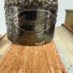 Puede incluir: Un servilletero plateado con detalles florales en relieve y el nombre "Anna May" en un &oacute;valo. Descansa sobre una superficie de madera envejecida, lo que sugiere un art&iacute;culo antiguo o vintage.