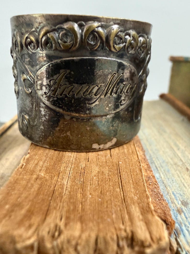 Puede incluir: Un servilletero plateado con detalles florales en relieve y el nombre "Anna May" en un &oacute;valo. Descansa sobre una superficie de madera envejecida, lo que sugiere un art&iacute;culo antiguo o vintage.