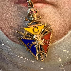 May include: A gold-toned metal pendant featuring a skull with red gemstone eyes above a triangular emblem. The triangle is divided into yellow, blue, and red sections with the letters C, E, and B, and features a second skull with crossbones.