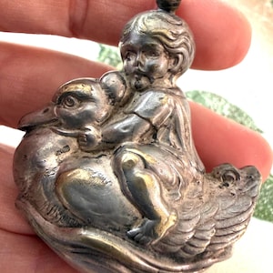 May include: A silver-toned metal pendant featuring a sculpted figure of a child riding on the back of a large bird, likely a swan or goose. The piece has a metallic finish with darkened recessed areas and a small loop at the top for attachment.