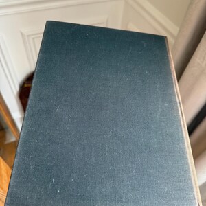 Puede incluir: Un libro de tapa dura con una cubierta de color verde azulado oscuro. La cubierta del libro tiene una apariencia texturizada y est&aacute; ligeramente inclinada, revelando el borde de las p&aacute;ginas. El libro se sostiene frente a un fondo neutro.