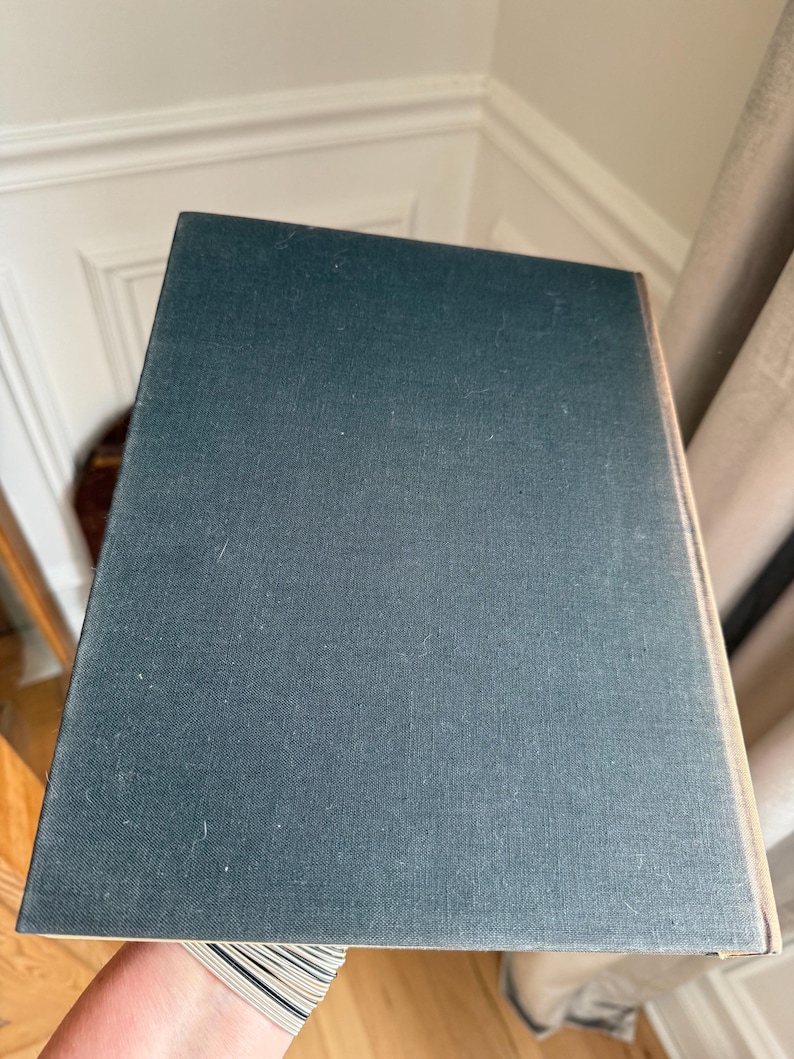 Puede incluir: Un libro de tapa dura con una cubierta de color verde azulado oscuro. La cubierta del libro tiene una apariencia texturizada y est&aacute; ligeramente inclinada, revelando el borde de las p&aacute;ginas. El libro se sostiene frente a un fondo neutro.