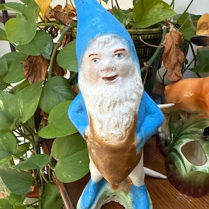 May include: A concrete garden gnome statue with a blue hat and boots, a white beard, and a brown tunic. The gnome has its hands on its hips and is standing on a green base. The statue is placed on a wooden surface.