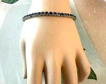 May include: A silver-toned tennis bracelet featuring a continuous row of small, dark, round-cut stones set in individual square metal links. The bracelet has a flexible design and a metallic finish, showcasing a uniform arrangement of dark gemstones.