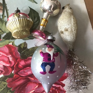May include: Four vintage glass Christmas ornaments. One is a silver ball with a pink base, one is a white bird with a silver tinsel tail, one is a silver cupcake with a green and gold design, and one is a silver teardrop with a painted gnome wearing a purple hat and blue pants.