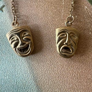 May include: Two antique gold-toned theater mask pendants, one smiling and one frowning, each suspended from a delicate chain. The pendants are detailed with facial features and are likely vintage costume jewelry.
