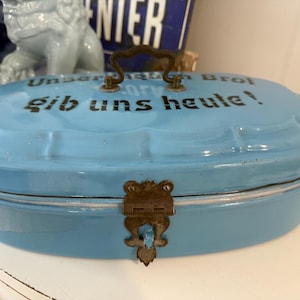 May include: A light blue, vintage enamel bread box with a handle and a decorative clasp. The lid features the German phrase "Unser täglich Brot gib uns heute!" in black lettering. A charming kitchen decor item.