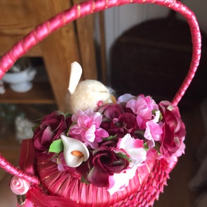 Puede incluir: Una cesta tejida rosa con un asa de cinta de sat&eacute;n rosa. La cesta est&aacute; llena de flores artificiales rosas y burdeos y un conejito blanco.