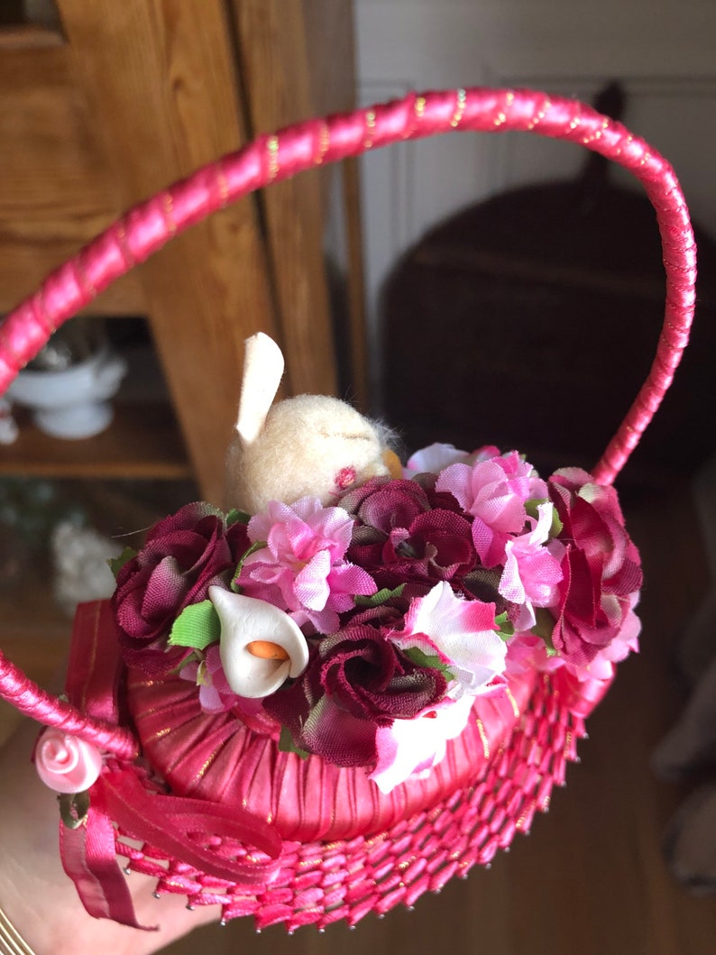 Puede incluir: Una cesta tejida rosa con un asa de cinta de sat&eacute;n rosa. La cesta est&aacute; llena de flores artificiales rosas y burdeos y un conejito blanco.