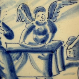 May include: Blue and white ceramic plate with a hand-painted scene. The image depicts a figure with wings, possibly an angel, seated at a table. Other figures and objects are also visible, all rendered in shades of blue.