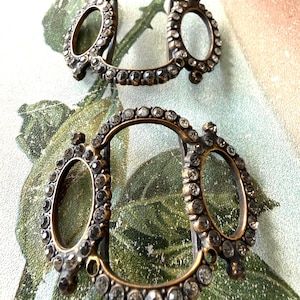 May include: Two antique bronze-toned buckles with oval frames and smaller oval accents, each encrusted with small, clear rhinestones. The buckles are displayed on a floral patterned background.